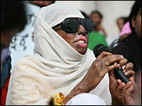 Haseena Hussain holds microphone, wearing dark glasses and covered by shawl